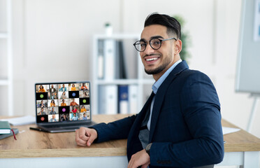 Poster - A young middle eastern businessman wearing glasses sits at his desk in an office, smiling as he looks away from his laptop. The laptop screen displays a video conference with many participants