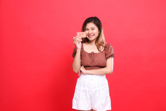 Asian woman smiles cheerfully with her arms outstretched, holding a debit credit card from the side, wearing a brown blouse, red background. for transaction, technology and advertising concepts
