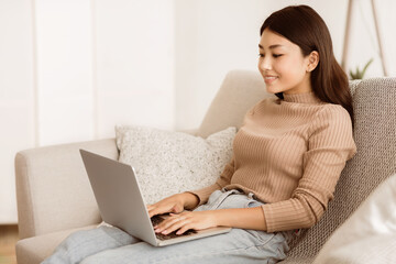 Poster - A young woman sits on a couch in a living room, working on her laptop computer. She is smiling and appears to be focused on her work.