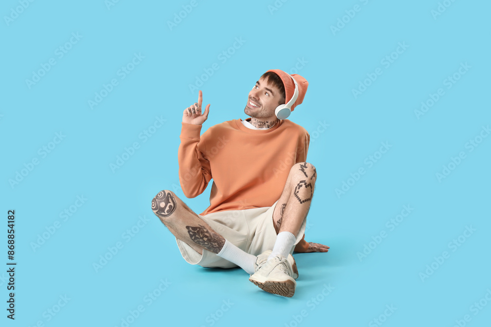 Young tattooed man in headphones pointing at something on blue background