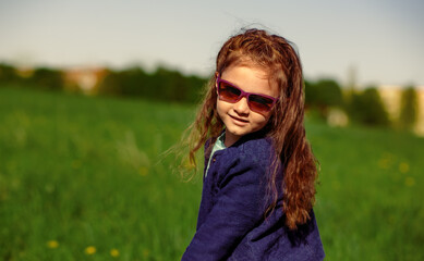 Wall Mural - Enjoying kid girl with beautiful long curly hairstyle walking on summer blue sky and green grass outdoors background in fashion violet sunglasses. Lifestyle