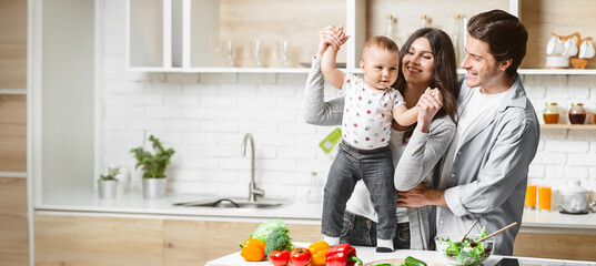 A happy family is playing in their modern kitchen while preparing food. The mother is holding up their young child, who is smiling and reaching out. Father is standing next to them, looking lovingly