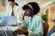 © Marko Geber - Stressed African American woman working on laptop in busy office environment