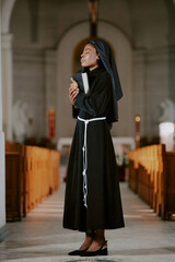 Wall Mural - Vertical long shot young African American nun holding Bible book standing in Catholic cathedral building with eyes closed