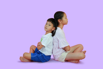 Wall Mural - Portrait of peaceful Asian young girl child and bored face little boy kid sitting back against each other isolated on purple studio background. Sister and brother relationship concept.