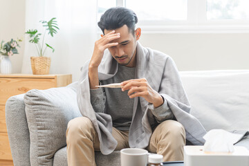 Sick, influenza asian young man, male have fever hand holding thermometer, touching forehead for check measure body temperature, illness while sitting rest on sofa at home. Health care on virus person