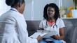 © The Little Hut - Young doctor woman holding report file with appointment and prescribe treatment to a female african american sick worried patient sitting on the couch in clinic during medical examinat : Generative AI