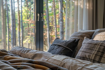 Wall Mural - A bed with grey plaid pillows and beige bedding, next to an open window overlooking the forest in autumn. The room is well-lit by natural sunlight filtering through sheer curtains.