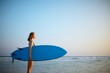 © kravtsov - A young surfer woman stands by the ocean with a blue surfboard before surfing at sunset