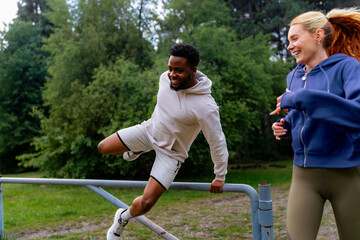 Wall Mural - Man jumping over a barrier while running, with a woman following behind. Both are laughing and having fun during their outdoor workout in the park.