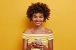 © JAYDESIGNZ - Photo of cheerful woman with curly hairstyle dressed striped top holding smartphone in hands smiling isolated on yellow color background