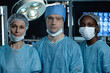 © Seventyfour - Waist up portrait of diverse surgical team of three people in operating room looking at camera in low light