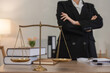 © wichayada - Confident Young Lawyer Standing in Modern Office with Scales of Justice and Legal Documents on Desk