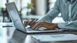 © naphat - A person in business attire is sitting at an office desk, typing on their laptop computer with one hand and holding papers or documents under the other arm. The background shows another individual's