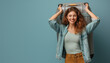 © gearstd - A joyful woman in a light green top and brown pants holds a suitcase behind her head with a smile, wearing a denim jacket, in front of a blue background, embodying excitement and readiness.