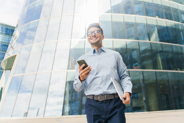 Smiling businessman with glasses holding a phone and tablet, standing near a modern glass building.