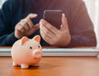© tete_escape - Save money, investment, digital banking accounting, business and financial concepts. Piggy bank on wood table near person hold smart mobile phone using for online deposit money in saving account.