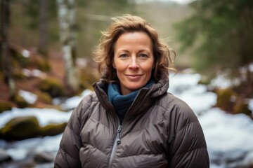 Sticker - Portrait of a cheerful woman in her 40s sporting a quilted insulated jacket while standing against tranquil forest stream