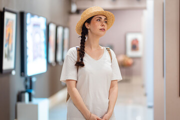 Wall Mural - Young Caucasian woman student in straw hat admiring paintings in the gallery. Concept of visit museum and cultural education