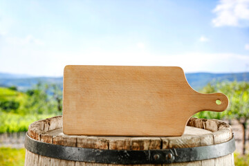 Wall Mural - Empty wooden cutting board on old wooden wine barrel with copy space and vineyard background.