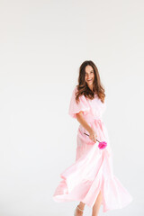 Wall Mural - Model wearing casual outfit. Look in trendy minimalistic style. Pink dress with slit, belt, puff sleeves. Sexy sensual happy woman. Positive woman turn around and hold pink flower on white background.