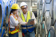 © chadchai - Senior engineer manager trains new employee within the metal sheet factory. Everyone wear safety vest and hardhat. Steel sheet roll stack are in the working area.