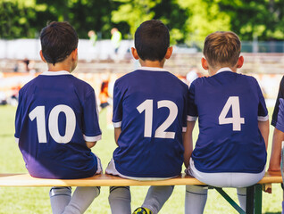 Wall Mural - Happy boys in a sports team. Kids in purple sporty clothes sitting in a team on a wooden bench. Boys in sports jersey shirts play a tournament game. Coaching little kids in sports