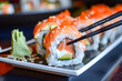 © mediahain.de - A close-up of sushi rolls with fresh fish, avocado, and rice, arranged neatly on a plate with a pair of chopsticks, against a blurred Japanese restaurant background