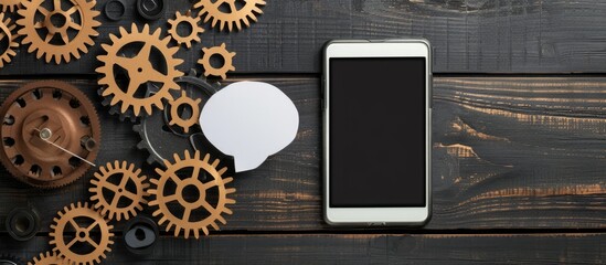 Poster - Image of a paper tablet or smartphone adorned with cogwheels and a speech bubble on a dark wooden backdrop. An abstract image with copy space.