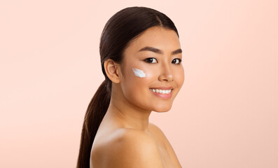 Wall Mural - This image shows Asian young woman with long, dark hair smiling at the camera. She is applying facial cream to her cheek. The background is a soft, light pink.