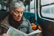 © yaqui_villegas - An elderly woman sits on a train with a backpack, focused intently on reading a map as she manages her travel plans, representing wisdom and adventure.