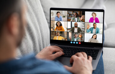 A man sits in a casual setting with a laptop on his lap, actively participating in a video conference with multiple participants. The screen displays a diverse group of people