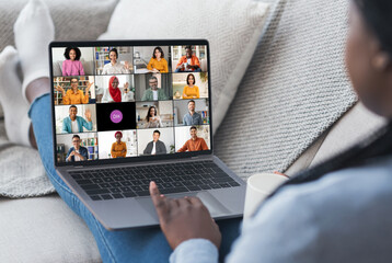 Poster - Black woman is sitting on a couch and participating in a video conference with colleagues. She is looking at the laptop screen, which shows a grid of video feeds of the other participants.