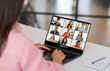 © Prostock-studio - A woman sits at a desk in a home office, looking at her laptop screen as she participates in a group video conference. The screen displays several participants in a grid format, webinar