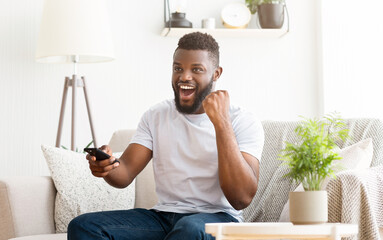 Wall Mural - A young Black man with a beard is sitting on a couch in his living room. He is watching television and holding a remote in his hand. He looks excited and is cheering.