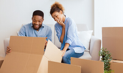 Wall Mural - Relocation. Happy Afro Spouses Unpacking Moving Boxes Sitting On Couch In New House. Selective Focus