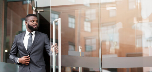 Wall Mural - Handsome confident african businessman in formal suit walking out of modern office center, copy space