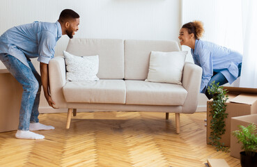 Wall Mural - Moving Concept. Cheerful Afro Spouses Placing Couch Furnishing Empty Room In New House After Relocation