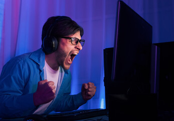 Wall Mural - An excited gamer wearing headphones sits in front of a computer screen, cheering with his fists in the air. The gamers face is lit by blue and purple lights in the background.