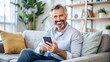 © PETR BABKIN - Happy mature man holding mobile phone while sitting on sofa at home.