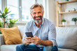 © PETR BABKIN - Happy mature man holding mobile phone while sitting on sofa at home.