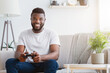 © Prostock-studio - African American man sits on a beige couch in a bright living room, holding a black video game controller in his hands. He is smiling and looking directly at the camera.