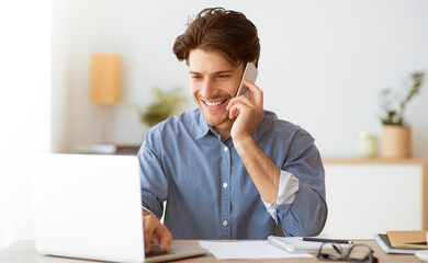Poster - A man is smiling and talking on a phone while working on a laptop. He is seated at a desk and has paperwork in front of him.