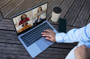 Poster - A person sits outdoors on a wooden patio, using a laptop to participate in a video conference. They are wearing a light blue shirt and white pants, top view