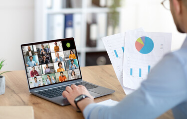 Wall Mural - A businessman sits at a desk in his office, using a laptop to participate in a virtual meeting with colleagues. The screen shows a grid of video calls with many diverse people