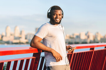 Wall Mural - Handsome african american sportsman smiling at camera, holding his phone, staying on bridge in sun light, copy space