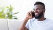 © Prostock-studio - A happy, smiling black man with a beard is talking on the phone while sitting on a white couch. He is wearing a light blue t-shirt and is looking off to the side, copy space
