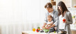 © Prostock-studio - A family of three is preparing food together in a kitchen. The father is holding their baby while they all help prepare vegetables. The mother is standing next to them, copy space