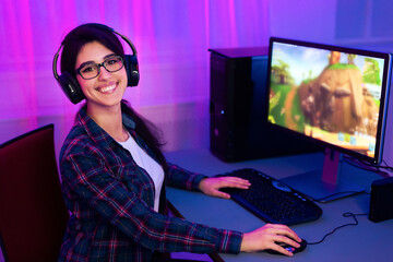 Poster - A young woman with long dark hair is smiling as she plays a computer game. She is wearing a plaid shirt and headphones. The computer monitor in the background shows an animated video game.