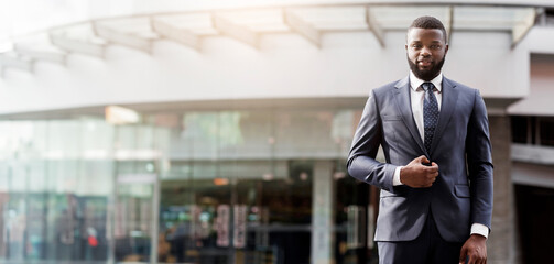 Wall Mural - Portrait of confident african american businessman in a stylish suit standing against office building looking at camera, copy space